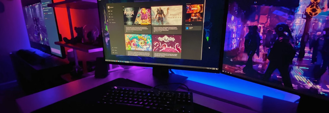 A gaming setup featuring multiple screens with vibrant game-related images illuminated by colorful LED lighting. The environment is dimly lit, highlighting the vivid displays on the monitors. A keyboard is visible on the desk beneath the central monitor.