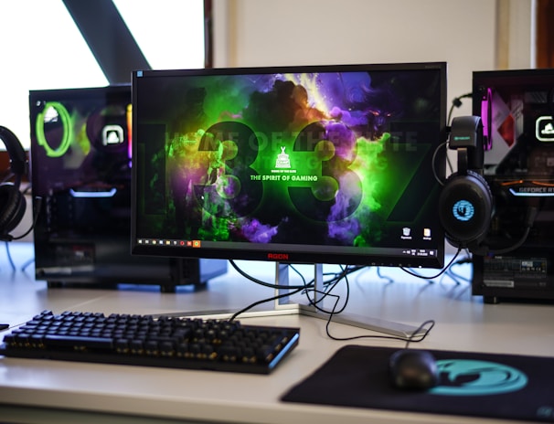 A gaming setup featuring a central monitor with a vibrant display showing 'The Spirit of Gaming'. On either side, there are gaming PCs with illuminated components and transparent cases. Headsets hang from the side of the monitor. The desk also has a black keyboard and mouse, and a branded mousepad.