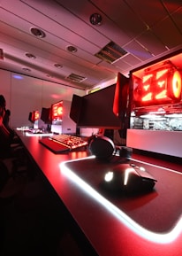A modern gaming setup featuring several high-end computers with illuminated red interior lighting. The room is equipped with gaming chairs exhibiting a sleek black and red design. Multiple monitors are arranged on a long desk, and peripheral devices like keyboards and mice are uniformly placed, reflecting an ambiance ideal for gaming or competitive play.