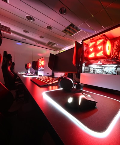 A modern gaming setup featuring several high-end computers with illuminated red interior lighting. The room is equipped with gaming chairs exhibiting a sleek black and red design. Multiple monitors are arranged on a long desk, and peripheral devices like keyboards and mice are uniformly placed, reflecting an ambiance ideal for gaming or competitive play.