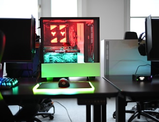 A modern gaming setup is displayed on a desk with multiple computer monitors and an open gaming PC tower. The tower is illuminated with red and green LED lights, showcasing a 'GeForce RTX' graphics card. A gaming mouse rests on a mousepad with glowing edges, and there is a keyboard with vibrant backlighting. The room appears well-lit with visible windows in the background.