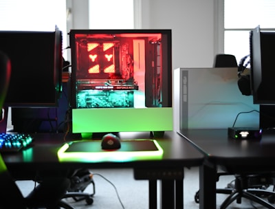 A modern gaming setup is displayed on a desk with multiple computer monitors and an open gaming PC tower. The tower is illuminated with red and green LED lights, showcasing a 'GeForce RTX' graphics card. A gaming mouse rests on a mousepad with glowing edges, and there is a keyboard with vibrant backlighting. The room appears well-lit with visible windows in the background.