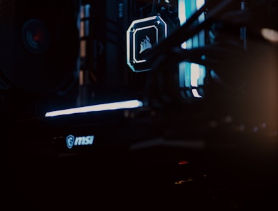 A close-up view of a computer gaming rig illuminated with blue LED lights. The interior shows components like cooling fans at the top, a glowing logo on a central component, and an MSI branded part at the bottom. The scene is dark, emphasizing the glow of the lights.