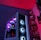 A gaming computer setup featuring a high-performance PC tower with multiple RGB fans lit in blue and purple hues. The case has a modern design with transparent panels showcasing internal components. In the background, shelves are stocked with various Ryzen and other gaming components under a vibrant red and blue ambient light.
