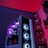 A gaming computer setup featuring a high-performance PC tower with multiple RGB fans lit in blue and purple hues. The case has a modern design with transparent panels showcasing internal components. In the background, shelves are stocked with various Ryzen and other gaming components under a vibrant red and blue ambient light.