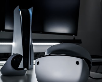 A sleek virtual reality headset sits on a reflective surface beside a gaming console and its controller. The background features a large dark monitor screen, creating a modern and futuristic setting. The lighting is subdued, highlighting the glossy surfaces of the electronic devices.