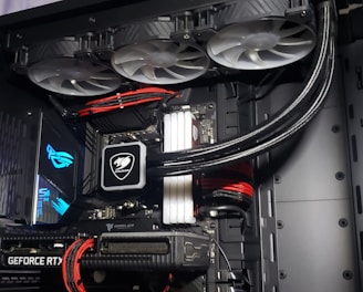 A high-performance gaming computer setup showing the interior components, including a GeForce RTX graphics card, cooling system with three fans, a motherboard, and visible wiring with red and black cables.