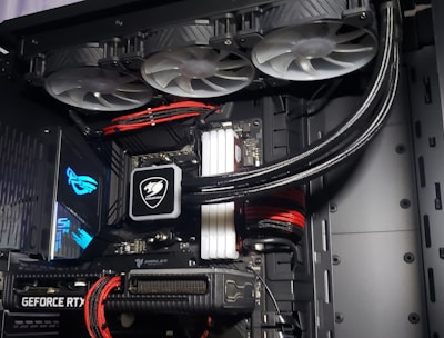 A high-performance gaming computer setup showing the interior components, including a GeForce RTX graphics card, cooling system with three fans, a motherboard, and visible wiring with red and black cables.