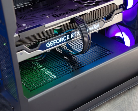 A close-up view of the interior of a computer casing showcasing a high-end graphics card with 'GeForce RTX' branding. The graphics card is secured in its slot, and several power cables are attached. The background reveals cooling fans illuminated with RGB lighting.