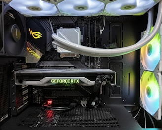 Inside view of a computer case showcasing a sophisticated gaming setup. Visible components include a GeForce RTX graphics card illuminated with green lighting, an ASUS motherboard, cooling fans with LED lighting, and a liquid cooling system with white tubes. Cables are neatly organized, and overall, the hardware suggests a high-performance machine.
