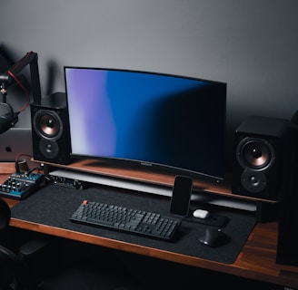 A modern gaming and streaming setup with a curved monitor placed centrally on a wooden desk. Surrounding the monitor are powerful speakers and a high-end PC tower with glowing fans. Various peripherals, including a mechanical keyboard, ergonomic mouse, and audio mixer, are neatly arranged. Overhead, a professional microphone and stand are ready for use, possibly for gaming commentary or podcasting.