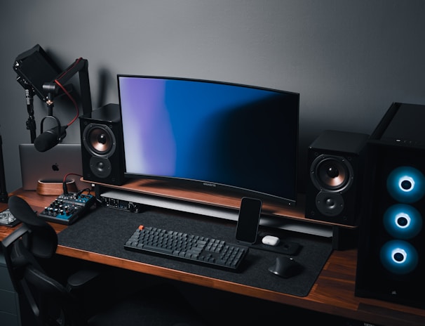A modern gaming and streaming setup with a curved monitor placed centrally on a wooden desk. Surrounding the monitor are powerful speakers and a high-end PC tower with glowing fans. Various peripherals, including a mechanical keyboard, ergonomic mouse, and audio mixer, are neatly arranged. Overhead, a professional microphone and stand are ready for use, possibly for gaming commentary or podcasting.